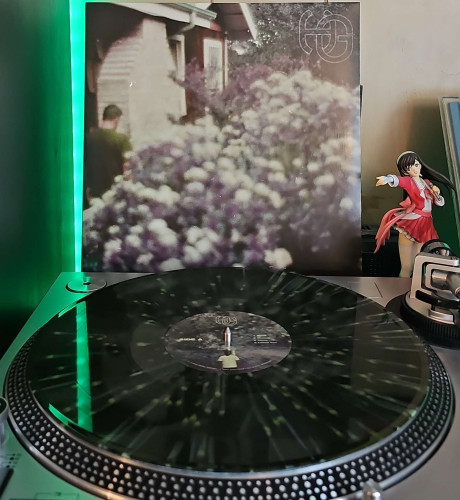 A Green Splatter vinyl record sits on a turntable. Behind the turntable, a vinyl album outer sleeve is displayed. The front cover shows the back of a man on the other side of a bush that has a bunch of flowers. To the right of the album cover is an anime figure of Yuki Morikawa singing in to a microphone and holding her arm out. 