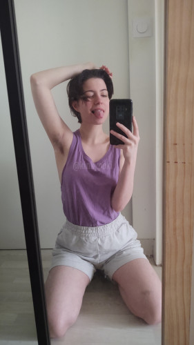 @kinkypoki kneeling in front of the mirror  wearing gray shorts and a purple tank top. They're sticking their tongue out and you can see their hairy armpit 
