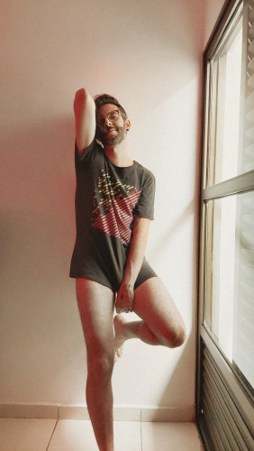 flux wearing a long t-shirt and no pants, the bottom of the shirt covering his cock but the fabric creates a visible bulge. He's leaning against a wall with one foot off the ground and sunlight is shining through a big window next to him.