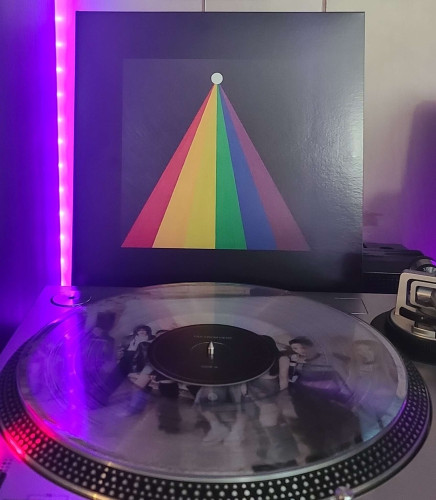 Image shows a turntable with a ckear vinyl record on the platter. Behind the turntable vinyl album outer sleeve is displayed. The front cover shows white orb near the top of a square, and there is a rainbow of colors stretching out from it.