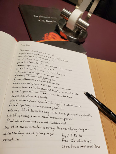 Handwritten transcription of the poem "The Sun" by A.F. Moritz from The Sentinel (House of Anansi Press) - an uncapped black pen rests on the notebook page and the poetry collection is in the background, with a cover including a bright orange sunburst, with a silver tape dispenser resting on it
