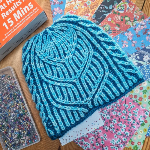 A two colour brioche hat in blues featuring a "whale tail" motif sits on top of origami papers, next to a box of sewing pins and a box of covid home tests 