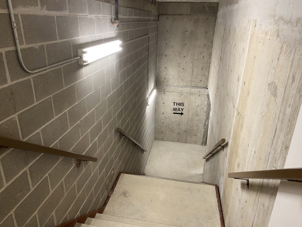 a concrete staircase in a concrete building. there is one fluorescent lamp and at the bottom of the staircase it reads “THIS WAY” with an arrow pointing to the right