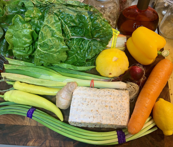 A cutting board with a lare bnch of chard, a yellow bell pepper, a yellow patty pan squash, 2 yellow banana peppers, a lemon, a large carrot, a bunch of scallions, 3 red young onions, a good-sized block of tempeh, a 3" section of thick ginger root, and a bundle of garlic stems.