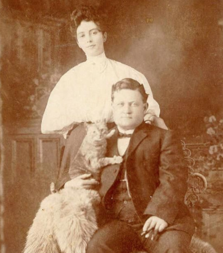 Sepia toned black and white photo of a stout white man in a three piece suit sitting on a chair draped with a light colored shaggy pelt of some kind. He has a shorthaired tabby cat (I believe it is orange) lovingly scooped to his side and the cat has its forepaws obligingly placed on his lapel. Behind these two stands a lovely young white woman in a voluminous Edwardian blouse, and all three look at the camera.