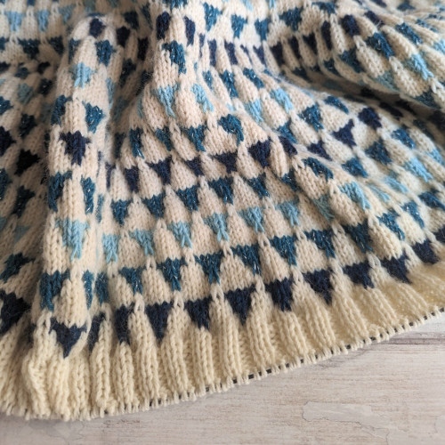 A knitting WIP on a white wood-effect background. The knitting is a white shawl with rows of blue triangles in different shades; the most frequent triangle colour is held with silver. The working edge has about an inch of 2x2 rib, with stitches on a circular needle.