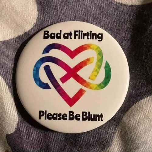 A pin-on button that says "Bad at flirting" at the top, and "Please be blunt" at the bottom, with the neurodivergent symbol interweaved with a similarly-multicolored heart.