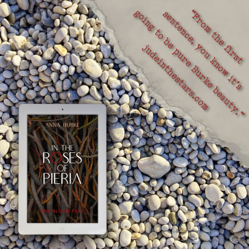 On a backdrop of pebbles, an iPad with the cover of In the Roses of Pieria by Anna Burke. In the top right corner of the image, a strip of torn paper with a quote: "From the first sentence, you know it's going to be pure Burke beauty." and a URL: judeinthestars.com.