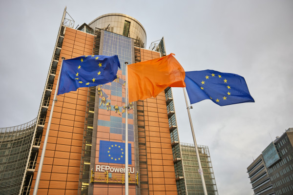Three flags flying in front of the European Commission headquarters in Brussels: two flags of Europe on the sides, and an orange flag in the middle.