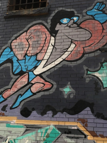 Image of a cartoon black man superhero leaping off into the sky.  He is the wearing a red outfit with blue boots, cape, and gloves. His sunglasses have blue lens with white star. This is the superhero from the School House Rock song  "Verbs--That's What's Happening"