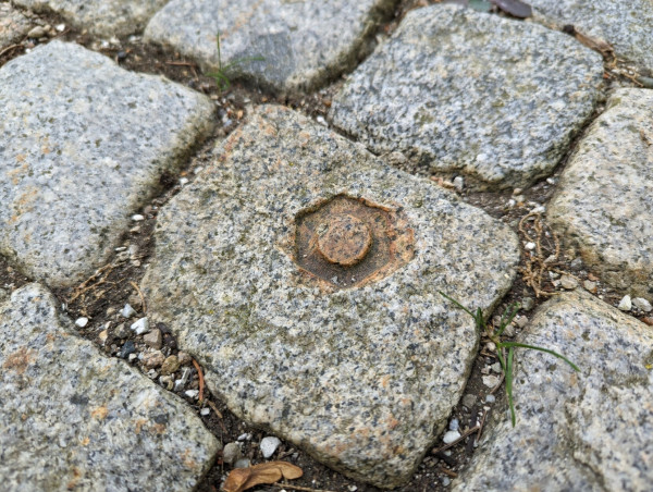 Foto von einem kleinen Granitpflasterstein. In der Mitte ein Abdruck von einer etwa Daumen-dicken  Mutter, etwas verrostet.
