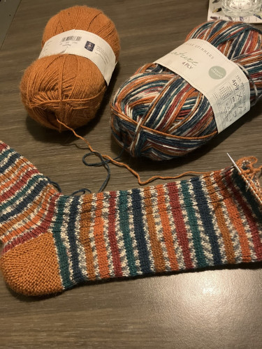 An almost finished knitted striped sock and two balls of yarn. The main parts of the sock are striped in rich blue, green, red, orange and brown-gold. The cuff and heel are brown-gold. 