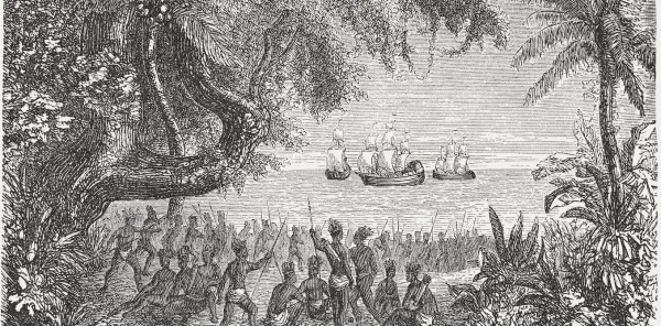 A black and white drawing shows a large crowd of Native American people wearing little clothing and holding spears, looking at boats in the water ahead of them. 