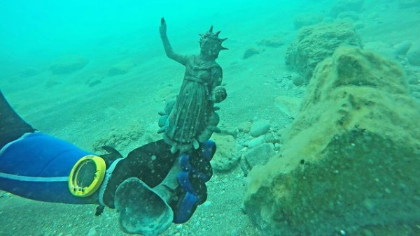 Underwater photo of a diver holding the statuette of the sun god Sol, dessed in a long robe and a cloak closed over his chest. He raises one arm, the other seems to hold a globe. He can be recognised by wearing his iconic sun ray crown.