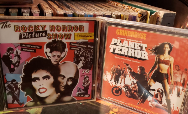 A photo of two Compact Disc jewel cases stood in front of a small rack full of other CDs. On the left is the score for the film "The Rocky Horror Picture Show." On the right is the the score for "Grindhouse: Planet Terror."