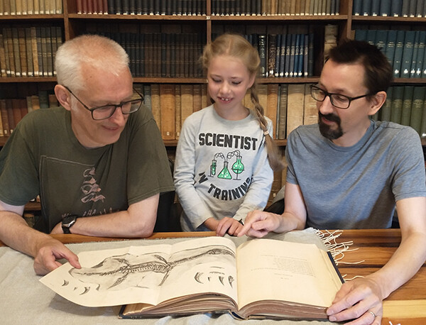 A man, Jonathan Emmett (left), a girl, Chloe, (middle), and a man (me), sitting at a table in a library, showing Chloe a book with an illustration of a plesiosaur skeleton.