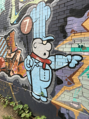 Street mural painted on the side of a brick building. Image of a white cartoon man in a blue engineering outfit (cartoon version) with a red scarf pointing. The emblem of the 7 train is just behind his blue conductor cap. Character is from the Schoolhouse Rock song "Conjunction Junction"