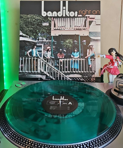 A translucent green vinyl record sits on a turntable. Behind the turntable, a vinyl album outer sleeve is displayed. The front cover shows the members of Banditos standing on a deck. 

To the right of the album cover is an anime figure of Yuki Morikawa singing in to a microphone and holding her arm out. 