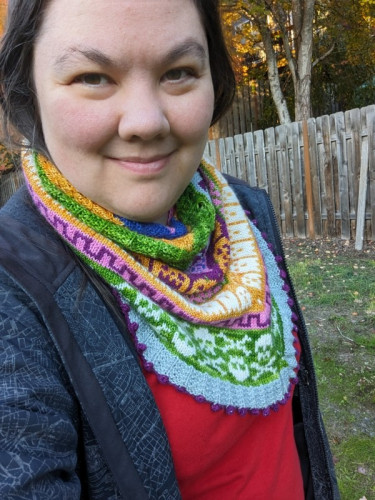 Terri, a mixed race woman, is wearing a knitted cowl with various Halloween motifs, with some fall foliage in the background.