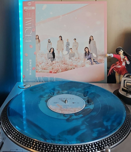 A transparent blue vinyl record sits on a turntable. Behind the turntable, a vinyl album outer sleeve is displayed. The front cover shows the 9 members of TWICE sitting and standing around in a field of flowers. 

To the right of the album cover is an anime figure of Yuki Morikawa singing in to a microphone and holding her arm out. 