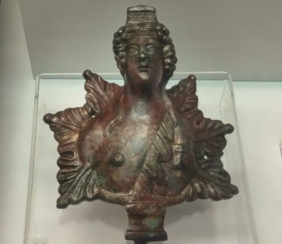 Bronze bust of the god Dionysos. He his shown with long hair flowing over his shoulders, one chest bare, the other covered in animal skin. Behind him, an arrangement of six leaves completes the decoration.