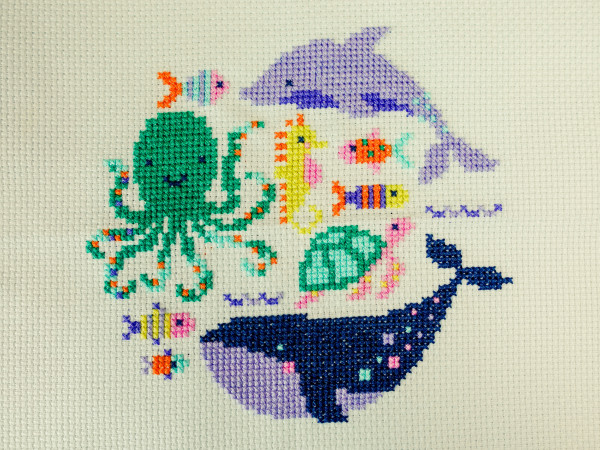Straight top-down view of a completed cross stitch project on light blue-green fabric. Various cute and smiling sea creatures including an octopus, dolphin, whale, seahorse, turtle and various fish. If the sea was really like this I would find it a lot less creepy!
