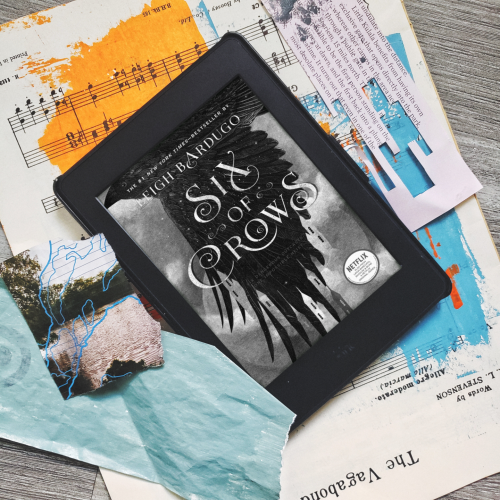 A ebook reader with the cover of the Six of Crows in the screen. The ebook reader is sitting on top of scraps of painted paper.