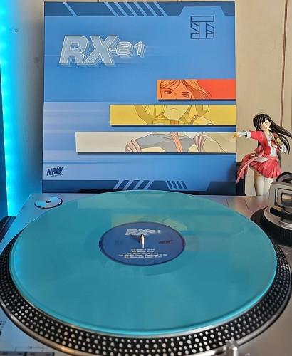 A blue vinyl record sits on a turntable. Behind the turntable, a vinyl album outer sleeve is displayed. The front cover shows an anime girl in a pilot outfit. Her image is broken in to 3 different colored panels.. 

To the right of the album cover is an anime figure of Yuki Morikawa singing in to a microphone and holding her arm out. 