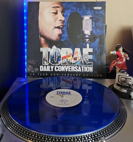 A translucent blue vinyl record sits on a turntable. Behind the turntable, a vinyl album outer sleeve is displayed. The front cover shows Torae rapping in to a studio microphone. 

To the right of the album cover is an anime figure of Yuki Morikawa singing in to a microphone and holding her arm out. 