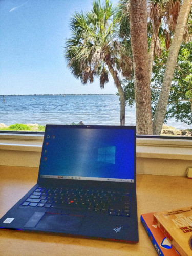 A laptop open on a wooden desk in front of a big window with a view palm trees and a river.