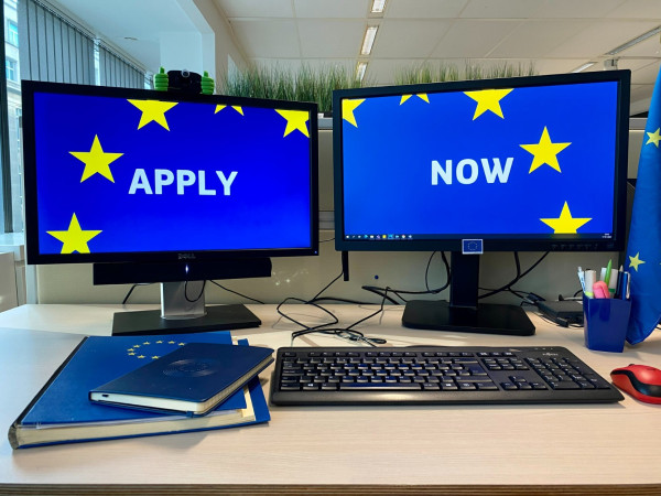 Two computer screens where you can read "Apply" on one of them lined with stars and "Now" on the other. You can also see some notebooks, a keyboard and a mouse on the desk. 