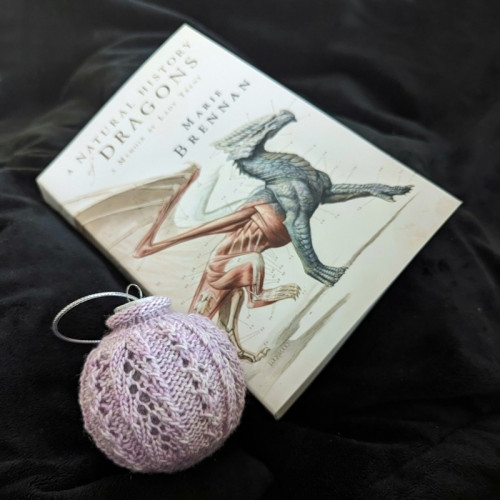 A lace knitted bauble sits next to a copy of A Natural History of Dragons. The book features an anatomical drawing of a dragon on the front.