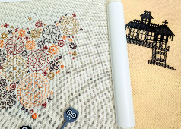 Two embroidery projects side by side. On the left, a delicate blackwork project in shades of yellow, brown and red. It is part of a world map made up of steampunk-style cogs. On the right, a solid black silhouette of a spooky house is being cross stitched on orange linen fabric.
