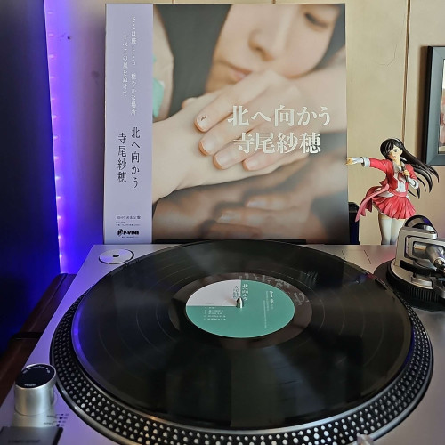 A vinyl record sits on a turntable. Behind the turntable, a vinyl album outer sleeve is displayed. The front cover shows Saho Terao resting her head on her arm. 