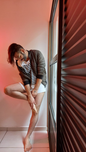 flux wearing a long t-shirt, a jacket and no pants, the bottom of the shirt is covering his cock. He's leaning against a wall, face looking down, natural light is shining through a big window next to him, and a soft diffused red light is coming from the other side.