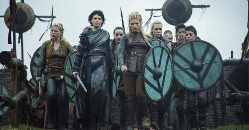 A group of warriors marches through an ancient settlement carrying swords and shields. They are all women.