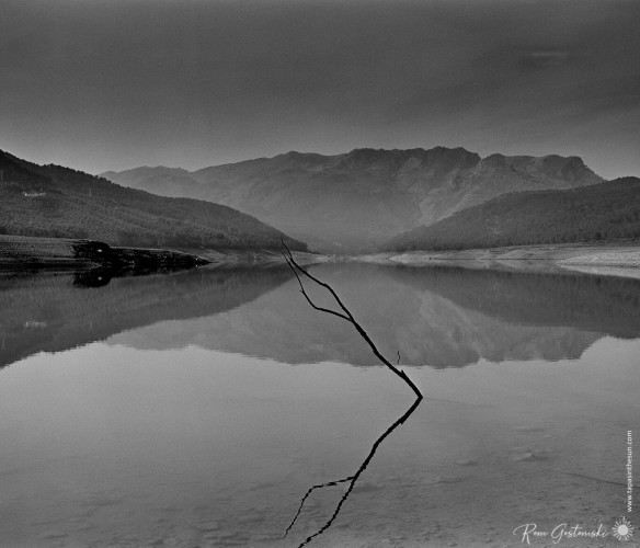 A black-and-white photo looking out along the reservoir towards the mountains. The water is still with clear reflections of a twig in the foreground and the mountains reflecting on the water's surface further away.