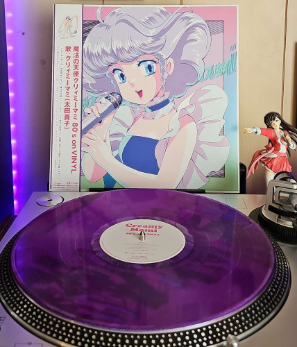 A translucent purple vinyl record sits on a turntable. Behind the turntable, a vinyl album outer sleeve is displayed. The front cover shows the anime character, Creamy Mami, singing in to a microphone. 

To the right of the album cover is an anime figure of Yuki Morikawa singing in to a microphone and holding her arm out. 