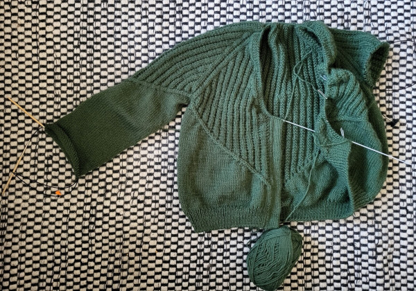 A green hand knitted cardigan is lying on the floor, part of the body is knitted in broken rib stitch and the rest in stockinette stitch. The cardigan isn't finished yet: one sleeve is still missing, the other one is halfway done. The double knit buttonband is only about 25cm done. One ball of green yarn lies next to the garment.