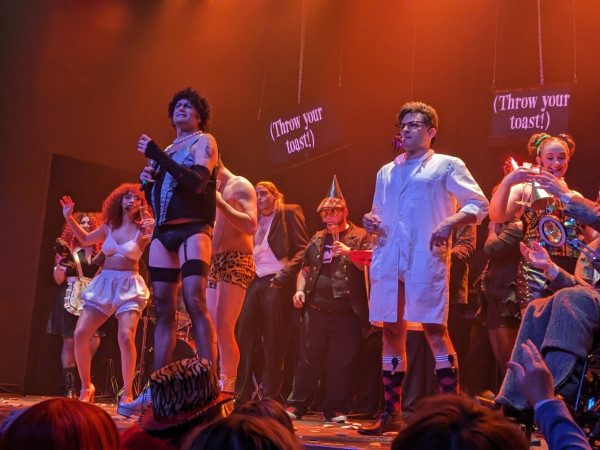 Most of the cast of "The Rocky Horror Show" performing on stage. Screens hanging in the background read "Throw your toast!"