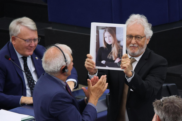 Héctor Abad Fasiolinse standing in the European Parliament hemicycle, holding a photo of Victoria Amelina, the young Ukrainian writer killed by a Russian missile. The other members of the Parliament and Commissioners surrounding him applaud. 