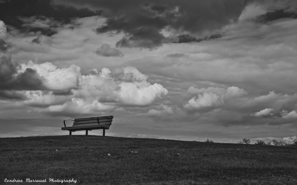A black-and-white photo of a bench at the top of a hill with a cloudy sky in the background.