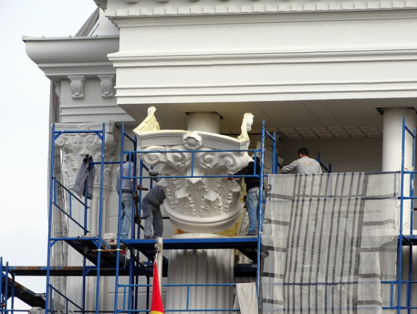 Four workers on a scaffolding are mounting a sheer white capitol made out of plaster onto a plain concrete column, making it appear like it was made in antiquity. The framed building surrounding the scene has almost completely been antiquified with decorative plaster molding.