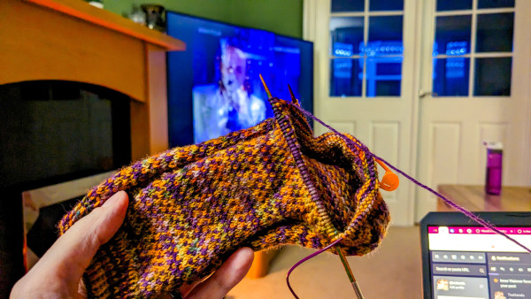 In-progress knitted Halloween sock in purple and orange yarn. Currently the gusset is under construction! In the background, Terrorvision are on the TV. You can just about see the corner of my laptop screen, where I'm keeping up with Mastodon's weekly Top of the Pops watchalong.