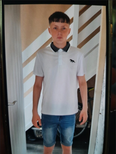 Photo of the missing young boy who is described in the post, in the photo he is wearing knee length jeans shorts, and a white polo neck shirt with a navy collar and a navy logo on the breast.