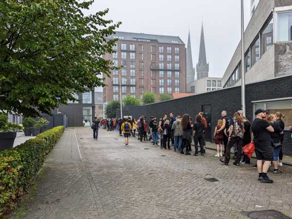 A long queue of people, mostly dressed in black, in a side alley next to a black brick wall. In the background there's a red building with the typical Dutch style, and the two towers of a church can be seen