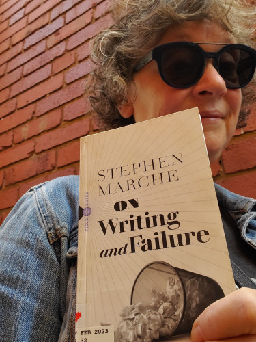 Me, standing against a red brick wall, with curly, gray hair, jean jacket and dark blue sunglasses, holding the book On Writing and Failure by Stephen Marche (Biblioasis), with an image of an overturned wastebasket on the cover