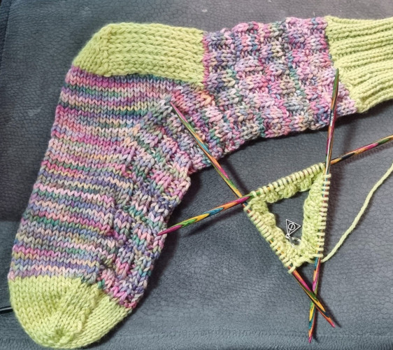 A knitted sock plus one started on needles.
Band, heel and toe in light green and main sock knitted in basketweave in colours from pinks, mauves, and greens.