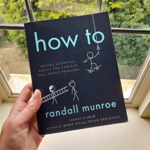 A white hand holding a copy of How To in front of a window. The black cover shows stick figures carrying a ladder and trying to change a lightbulb while balancing on drones.
