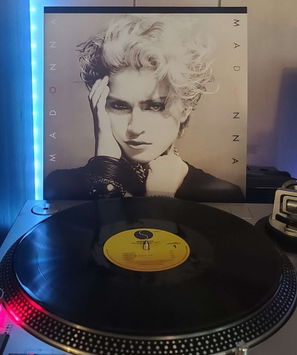 Image shows a turntable with a black vinyl record on the platter. Behind the turntable vinyl album outer sleeve is displayed. The front cover shows Madonna from the shoulders up. She has short hair styled up. She has her hand to her face and is looking at the camera. 